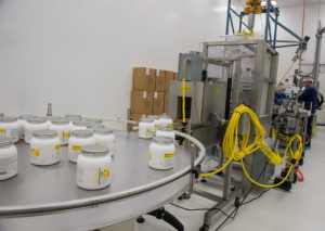 IQ Formulation packaging equipment along the wall with an employee off in the background on the right. The left foreground show the packaged products waiting on a round table attached to the equipment.