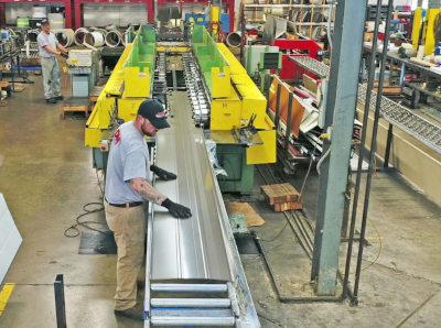 A worker in the foreground with his hands on a metal sheet on a conveyor. Machinery all around in a large room.