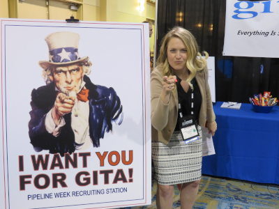 A woman at a conference, standing next to a sign with uncle same that says I want you for GITA!. The woman is doing that same forward look and pose with her hand pointing towards the camera, like the uncle Sam poster.