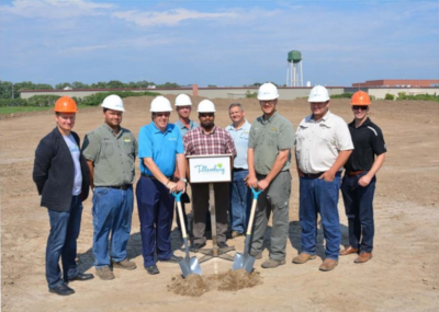 A Tillsonbirg breaking ground ceremony with 9 men in hard hats standing. Two men with shovels in the ground and a sign that reads Tillsonburg in the middle.